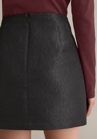 Zwarte mini-jurk met textuur, gekenmerkt door een reliëfpatroon, een verborgen rits aan de achterkant en een getailleerde pasvorm. Draagt bij een bordeauxrode top met lange mouwen.