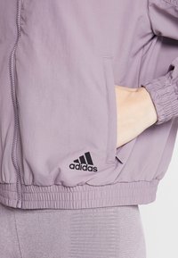 Ljuslila Adidas zip-up jacka med elastiska muddar och fåll. Har en sidoficka och svart logo på vänster bröst. Glatt material.