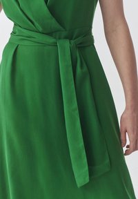 Rochie verde cu un material neted, design încrețit și un brâu legat în talie. Materialul are un finisaj mat și o culoare uniformă.