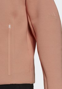 Manche et côté de sweat à capuche Adidas marron clair avec logo embossé sur le bras et poche verticale zippée portant l'étiquette Z.N.E.