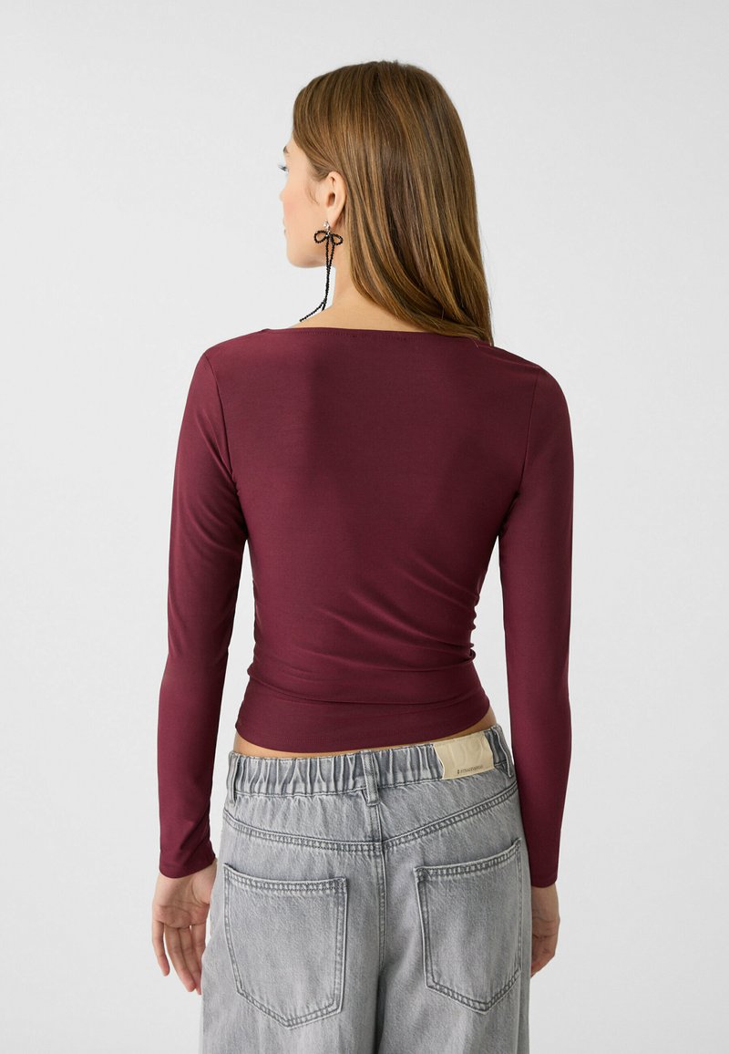 Donna con lunghi capelli castani che indossa un top attillato a maniche lunghe di color marrone scuro e jeans denim grigi chiari, girata di spalle su uno sfondo semplice.