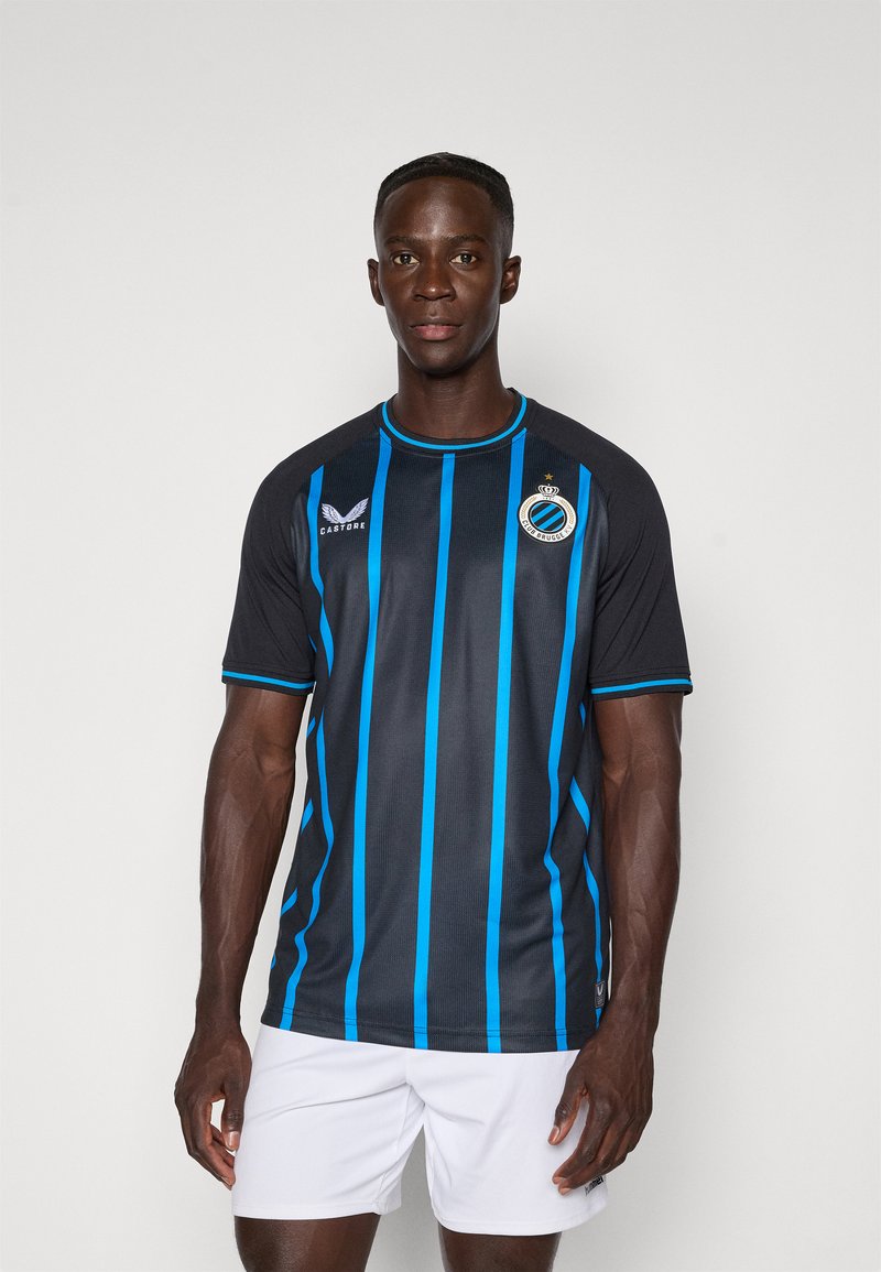 Atleta masculino em pé, vestindo uma camisola de futebol do Club Brugge em preto e azul listrado e calças brancas, contra um fundo claro.