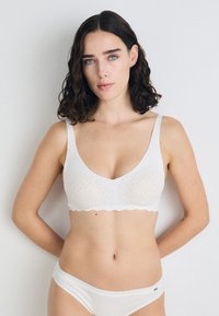 Bralette blanche avec un bord festonné, fabriquée en tissu texturé. Elle dispose de larges bretelles et d'un décolleté ras du cou. Associée à des culottes blanches assorties.