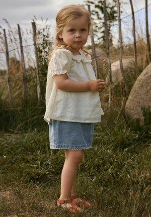 Mlada djevojka plave kose u bijeloj bluzi i traper suknji drži suhu stabljiku trave i stoji na travnatom tlu blizu drvenog ograde i stijena.