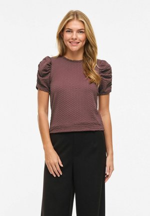 Femme souriante portant un haut violet à manches bouffantes texturé et un pantalon noir, debout devant un fond blanc uni.