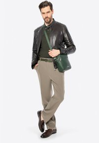 Borsa a tracolla verde in pelle con una finitura texture, indossata con una giacca in pelle nera, un maglione verde, pantaloni beige e scarpe marroni.