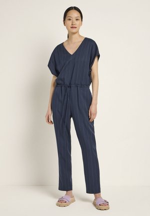 Dunkelblauer Jumpsuit mit weißen Streifen, V-Ausschnitt, kurzen Ärmeln und gebundenem Taillengürtel. Getragen mit helllila Sandalen und neutralem Hintergrund.