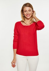 Roter Strickpullover mit Rundhalsausschnitt, langen gerippten Ärmeln und lockerer Passform, kombiniert mit weißen Hosen. Glatte Textur, mittelschweres Material.
