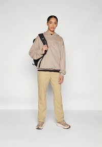 Béžová mikina s kapucňou a sťahovacími šnúrkami, tmavá vrstva pod ňou, svetlo khakové nohavice a béžové tenisky. Čierny batoh prehodený cez rameno.