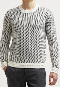 Pull blanc en maille avec motif géométrique noir, col rond, poignets et ourlet côtelés, présentant un design de tissu texturé.