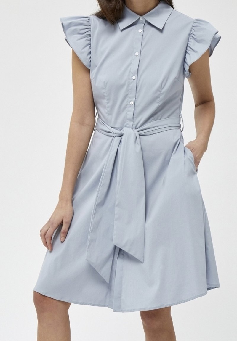 Robe sans manches bleu clair avec des manches courtes à volants, boutons sur le devant et une large ceinture en tissu nouée à la taille, portée par une personne debout.