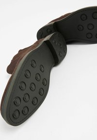 Zapato de ante marrón con una suela de goma negra que presenta patrones de agarre circulares. La suela muestra textura y tiene una forma elegante y redondeada.