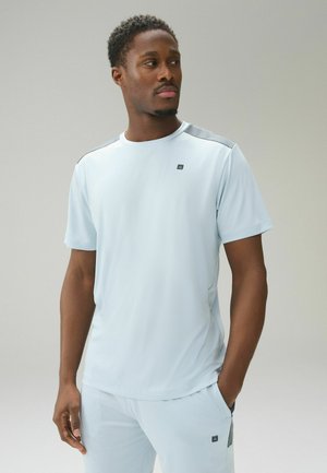 Mann trägt ein hellblaues kurzärmeliges Sportshirt und passende Hose, steht mit einer Hand in der Tasche und schaut zur Seite.