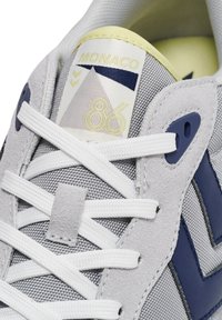 Szary sneaker wykonany z teksturowanej skóry zamszowej i siateczkowych materiałów, z białymi sznurówkami, niebieskimi akcentami oraz metką z logo o geometrycznym wzorze.