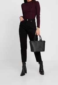 Kvinna i mörklila ribbad långärmad topp, högmidjade svarta jeans, svarta klackboots, hållande en svart läderhandväska i handtagen.