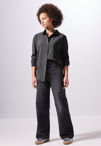 Camicetta verde con maniche arrotolate, abbinata a jeans scuri con decorazioni di borchie bianche e sneakers argentate.