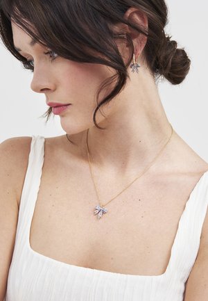 Kobieta o ciemnych włosach w białym topie bez rękawów, nosząca złoty naszyjnik i pasujące kolczyki z fioletowymi, kwiatowymi wisiorkami w kształcie kokardek.