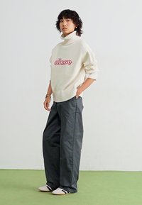 Hvid strikket turtleneck sweater med rødt "ellasse" logo, parret med løse grå bukser og sort-hvide stribede sko.