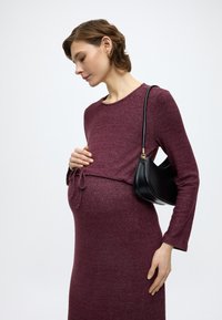 Sukienka ciążowa w głębokim bordowym kolorze z długimi rękawami i wiązaniem w talii, zestawiona z małą czarną skórzaną torebką na ramię.