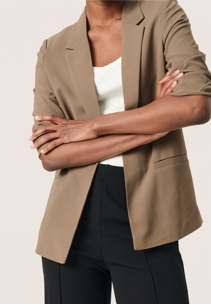 Jasnobrązowa marynarka o dopasowanym kroju, z klapami z wycięciem i dwoma bocznymi kieszeniami, noszona na białej prążkowanej koszuli i czarnych spodniach.