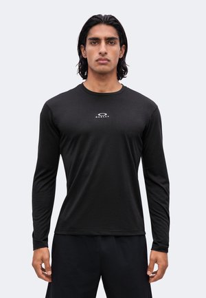 Junger Mann mit mittlerem Hauttyp und dunklen Haaren, der ein schwarzes langärmliges Oakley-Shirt und schwarze Hosen trägt, vor einem schlichten hellen Hintergrund stehend.