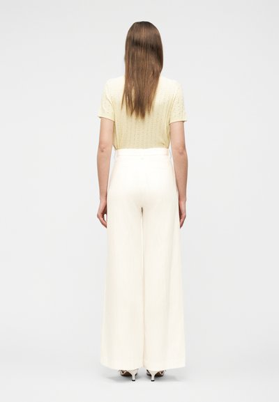 Γυναίκα που στέκεται με την πλάτη, φοράει ανοιχτό κίτρινο κοντομάνικο τοπ, ψηλόμεσο παντελόνι με φαρδιά μπατζάκια σε λευκό χρώμα και λευκά παπούτσια με τακούνι.