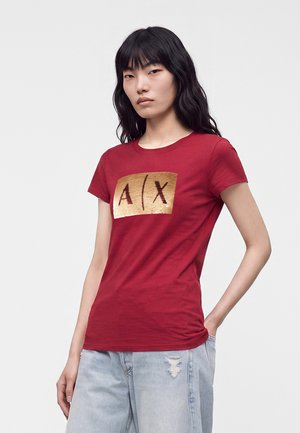 Young woman with long black hair wearing a red t-shirt with a gold sequined A|X logo and light blue ripped jeans.