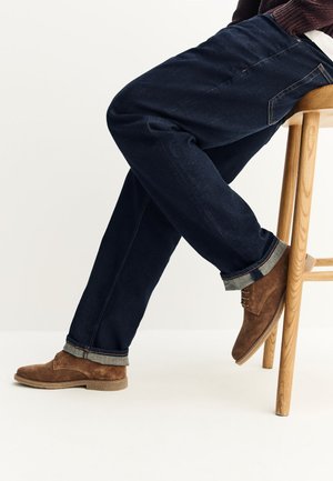 Person sitting on wooden stool wearing dark blue jeans with rolled cuffs and brown suede lace-up shoes on white background.