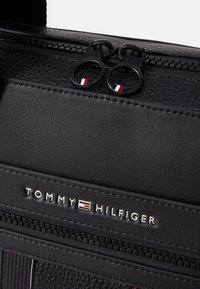 Zwarte synthetische tas met een gestructureerde afwerking, voorzien van een dubbele rits met ronde trekkers en een metalen logo plaat met de tekst "TOMMY HILFIGER."