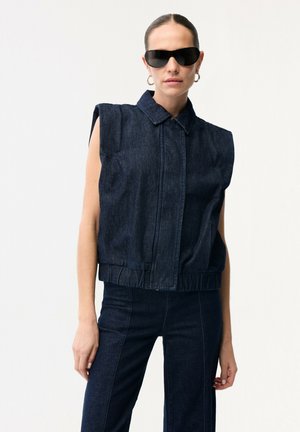 Donna che indossa un top smanicato in denim scuro con spalle imbottite, pantaloni in denim scuro abbinati, occhiali da sole neri e orecchini a cerchio, in piedi su sfondo bianco.