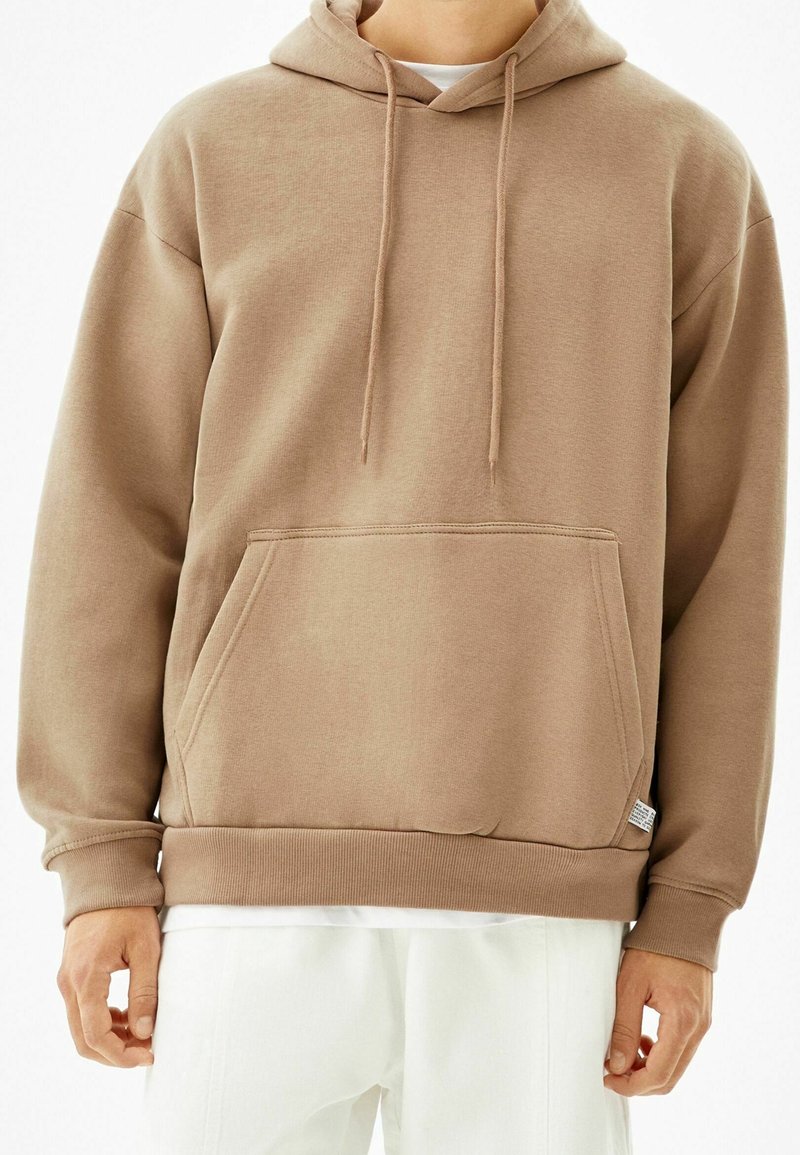 Personne portant un sweat à capuche beige uni avec une poche avant et des cordons, associé à un pantalon blanc ample, sur un fond blanc.