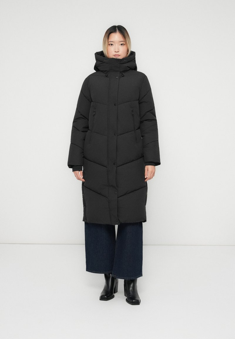 Kvinde iført en lang sort teddy-pufferjakke med hætte, mørke vide jeans og sorte ankelstøvler med hæl, stående mod en ensfarvet hvid baggrund.