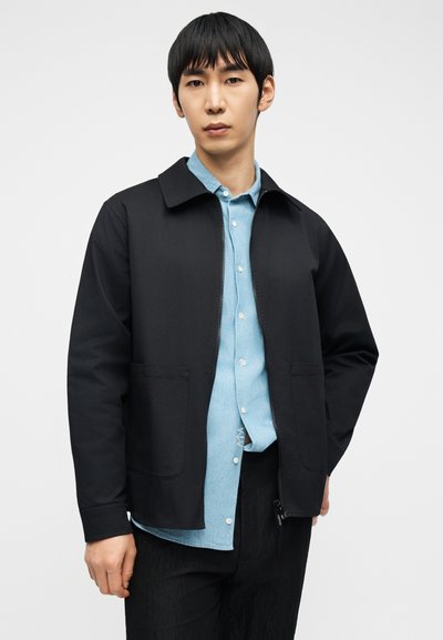 Νέος άνδρας που φοράει μαύρο μπουφάν με φερμουάρ πάνω από ανοιχτό μπλε πουκάμισο με κουμπιά και μαύρο παντελόνι, στέκεται μπροστά σε απλό λευκό φόντο.