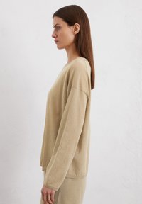 Haut beige léger à manches longues avec col rond et épaules tombantes, affichant une texture douce et une coupe décontractée.