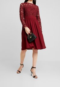 Burgundské šaty s krajkovými dlouhými rukávy a těsným korzetem, splývavou sukní, doplněné malou černou kabelkou a černými sandály na podpatku.