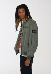 Giacca bomber verde oliva con zip frontale, polsini a coste e tasca laterale; presenta toppe ricamate e una texture liscia e lucida.