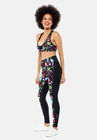 Sport-BH und Leggings-Set mit bunten geometrischen Mustern in Grün, Pink und Blau auf schwarzem Stoff; glatte Textur. Weiße Sneakers.