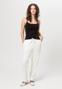 Zwarte tanktop, witte taps toelopende broek met trekkoord, en beige schoenen met zwarte accenten. Krullend haar, handen in de zakken, staande pose.