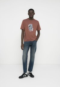 Μπλούζα σε χρώμα σκουριάς με γραφικό σχέδιο, συνδυασμένη με μπλε τζιν τζιν και μαύρα αθλητικά παπούτσια με λευκές ρίγες. Απλός σχεδιασμός, κανονική εφαρμογή.