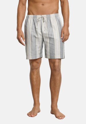 Hombre con pantalones cortos beige hasta la rodilla con rayas verticales grises y cintura ajustable con cordón, de pie descalzo frente a un fondo liso.