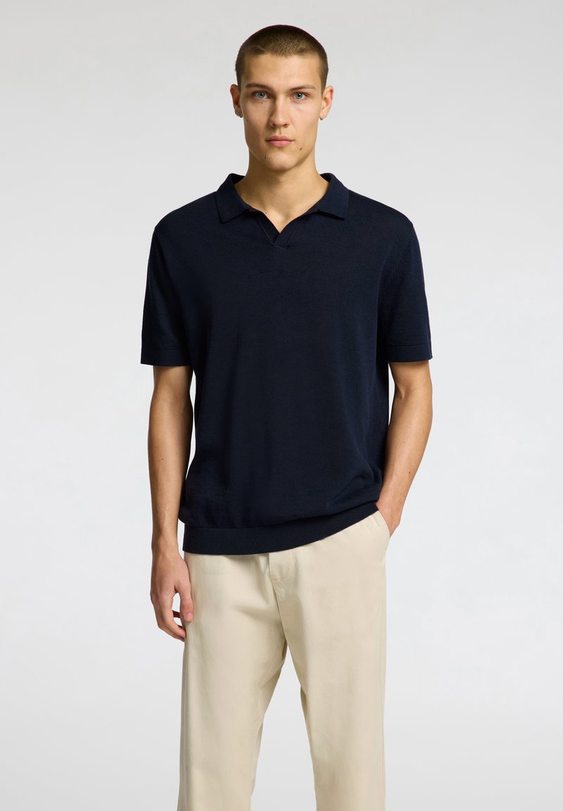 Navy short-sleeve polo made of textured fabric, featuring a collar and a relaxed fit, paired with light beige trousers.