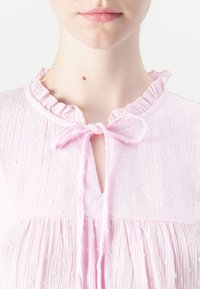 Blusa rosa chiaro con tessuto testurizzato, colletto arricciato e dettaglio con fiocco centrale. Presenta pieghe arricciate sotto il collo.