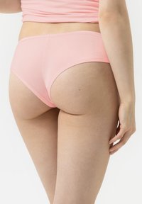 Vrouw in lichtroze ondergoed en bijpassende top, van achteren gezien, tegen een eenvoudige witte achtergrond.