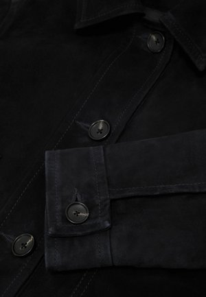 Primer plano de una chaqueta de ante negra que muestra el puño abotonado, los botones y la costura detallada en la tapeta frontal y el cuello.