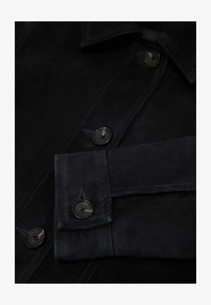 Közeli felvétel egy fekete velúr dzsekiről, amelyen gombos mandzsetta, gombok és részletes varrás látható az elején lévő záródáson és a galléron.