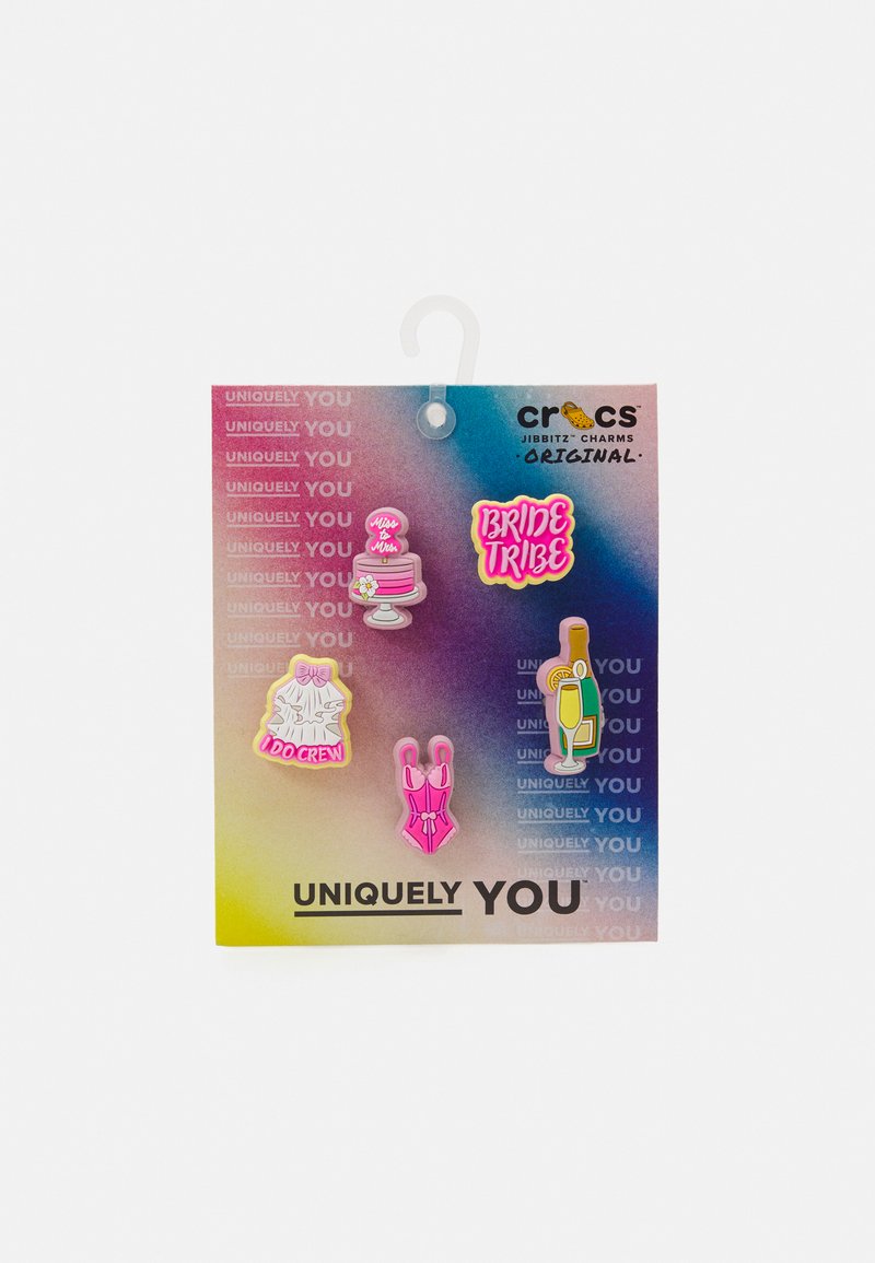As figuras Jibbitz da Crocs sobre um fundo em degradê incluem um bolo rosa, o texto "Bride Tribe", um vestido branco, um fato de banho rosa e uma garrafa de champanhe.