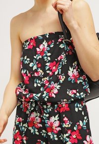 Jumpsuit bez ramínek černé barvy s květinovým vzorem v červené, růžové a tyrkysové barvě, s úzkým pasem, a osoba, která drží černou kabelku přes rameno.
