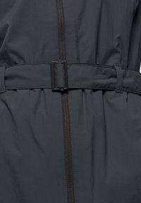 Giacca grigio scuro con zip frontale e cintura con fibbia rettangolare. La texture è liscia con una finitura leggermente lucida.