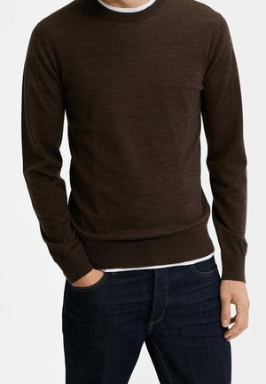 Brown knit sweater with a round neckline, long sleeves, and ribbed cuffs and hem. Worn over a white shirt, paired with dark blue jeans.
