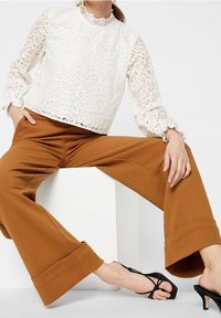 Blouse en dentelle blanche avec un col rond et des manchons volants, associée à un pantalon marron à jambes larges avec un ourlet plié, et des sandales noires à lanières.